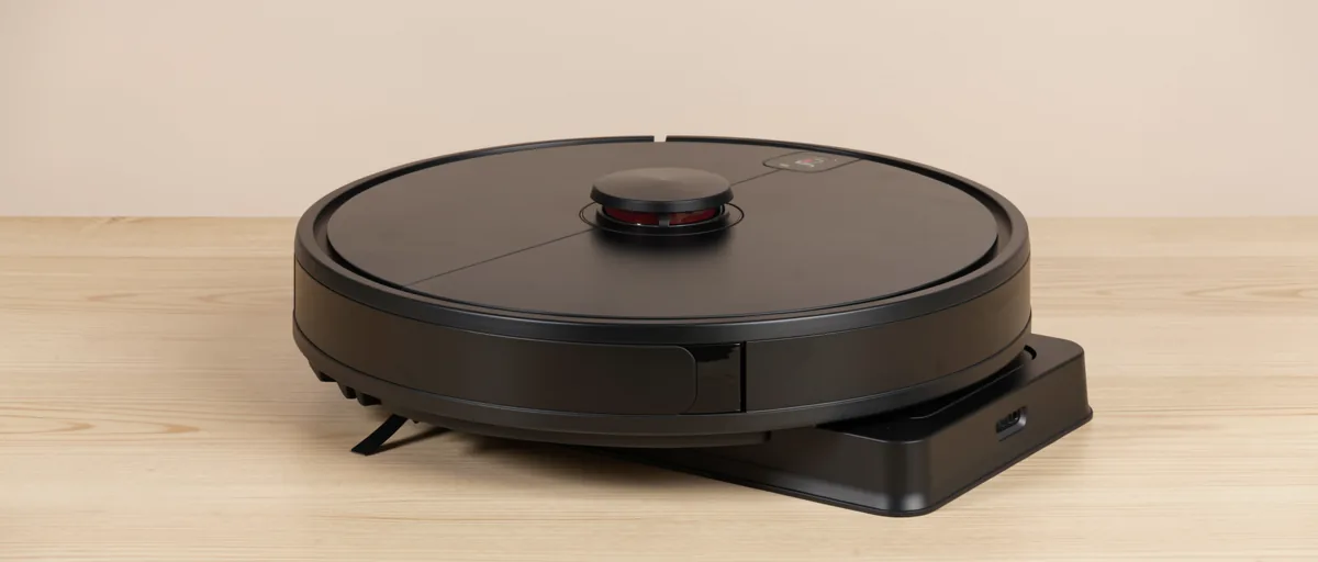 A round black robot vacuum cleaner resting on a light wooden floor next to its charging dock — illustration for Best Robot Vacuum Cleaner in 2026
